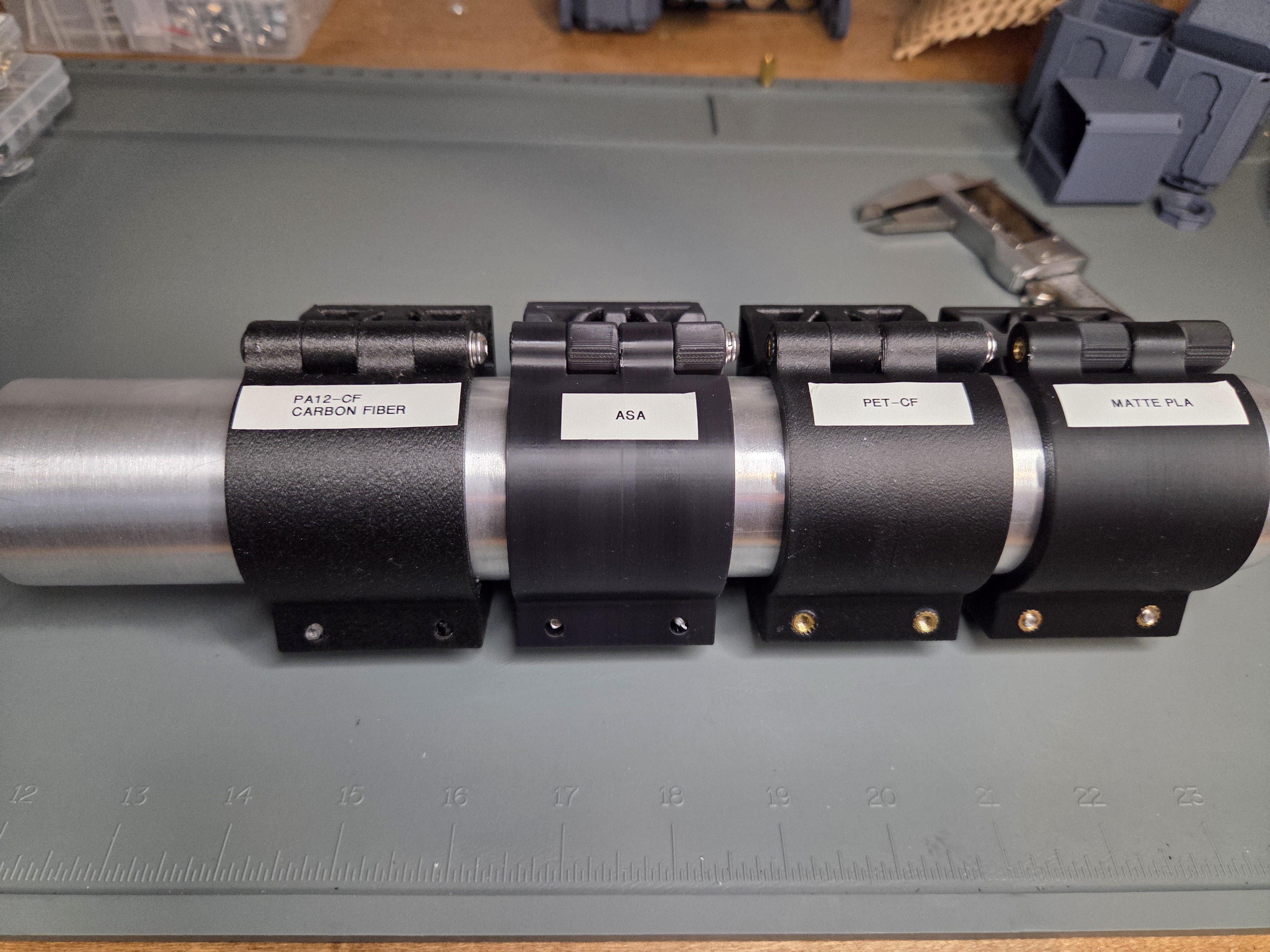 Side-by-side comparison of the same clamp body printed in PA12-CF, ASA, PET-CF, and matte PLA